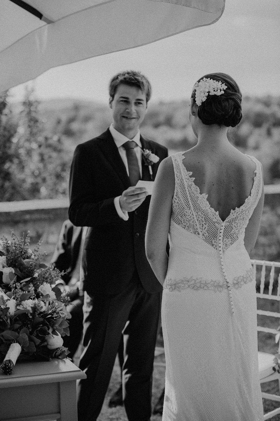 Black and white photo of smiling groom reading vows to bride in elegant backless lace wedding dress outdoors. Tuscany Villa Catignano