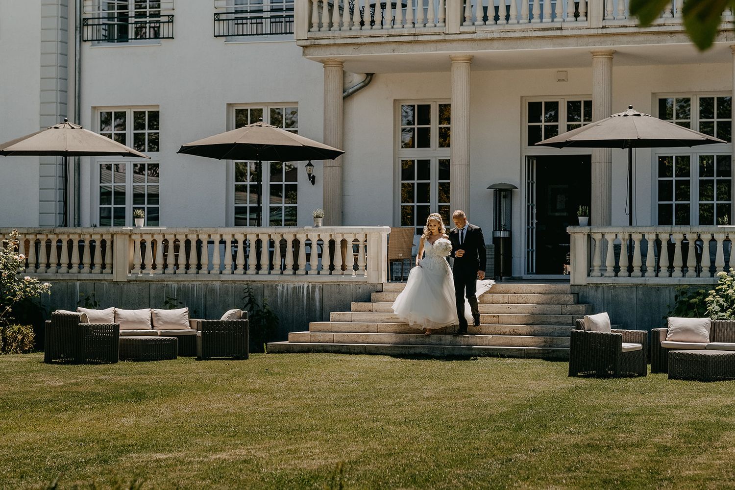 Bride and groom pose on steps of elegant white manor house surrounded by manicured garden lawn. Hideaway Starnberger See