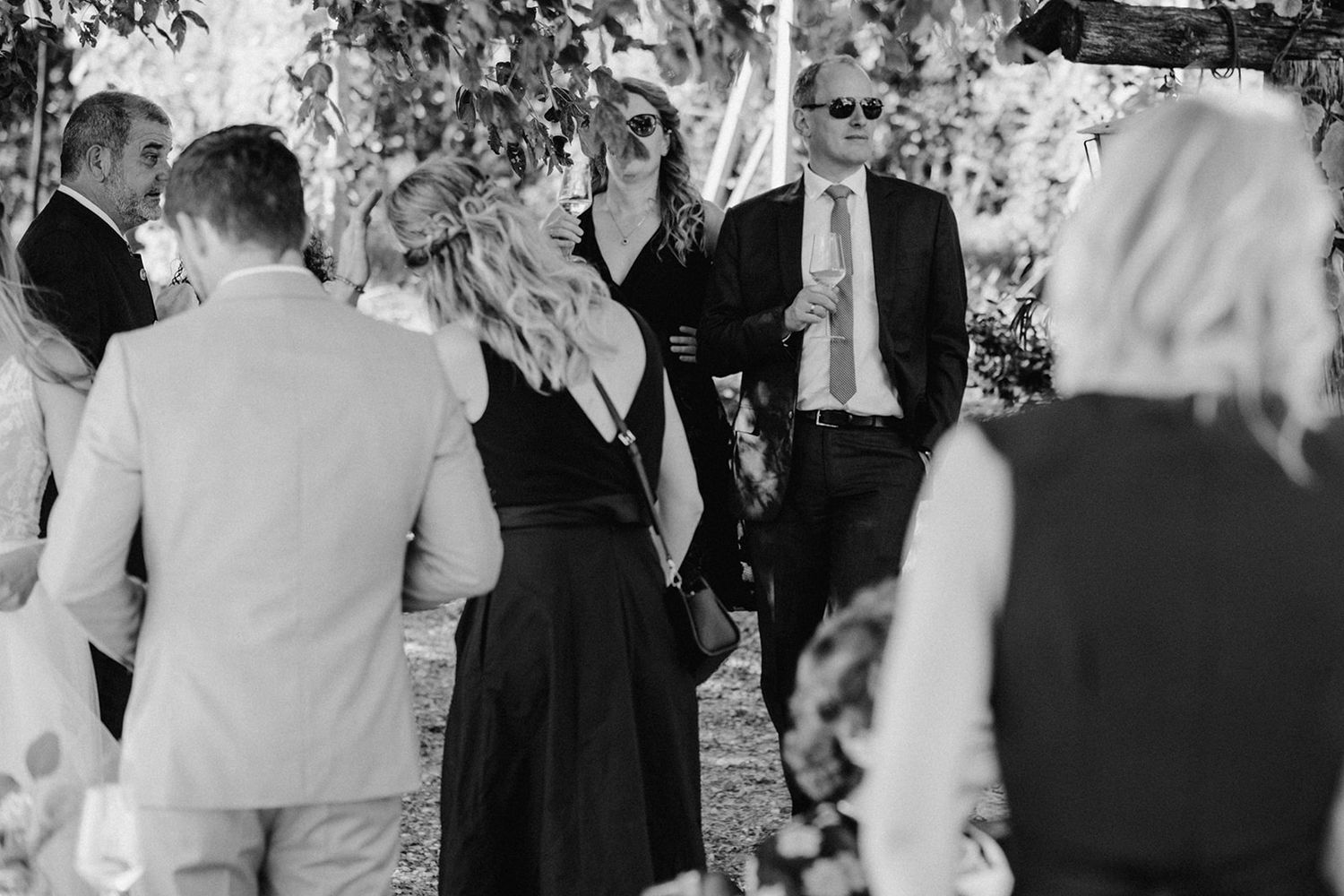 Wedding guests socializing outdoors under trees in black and white photography at a garden ceremony.