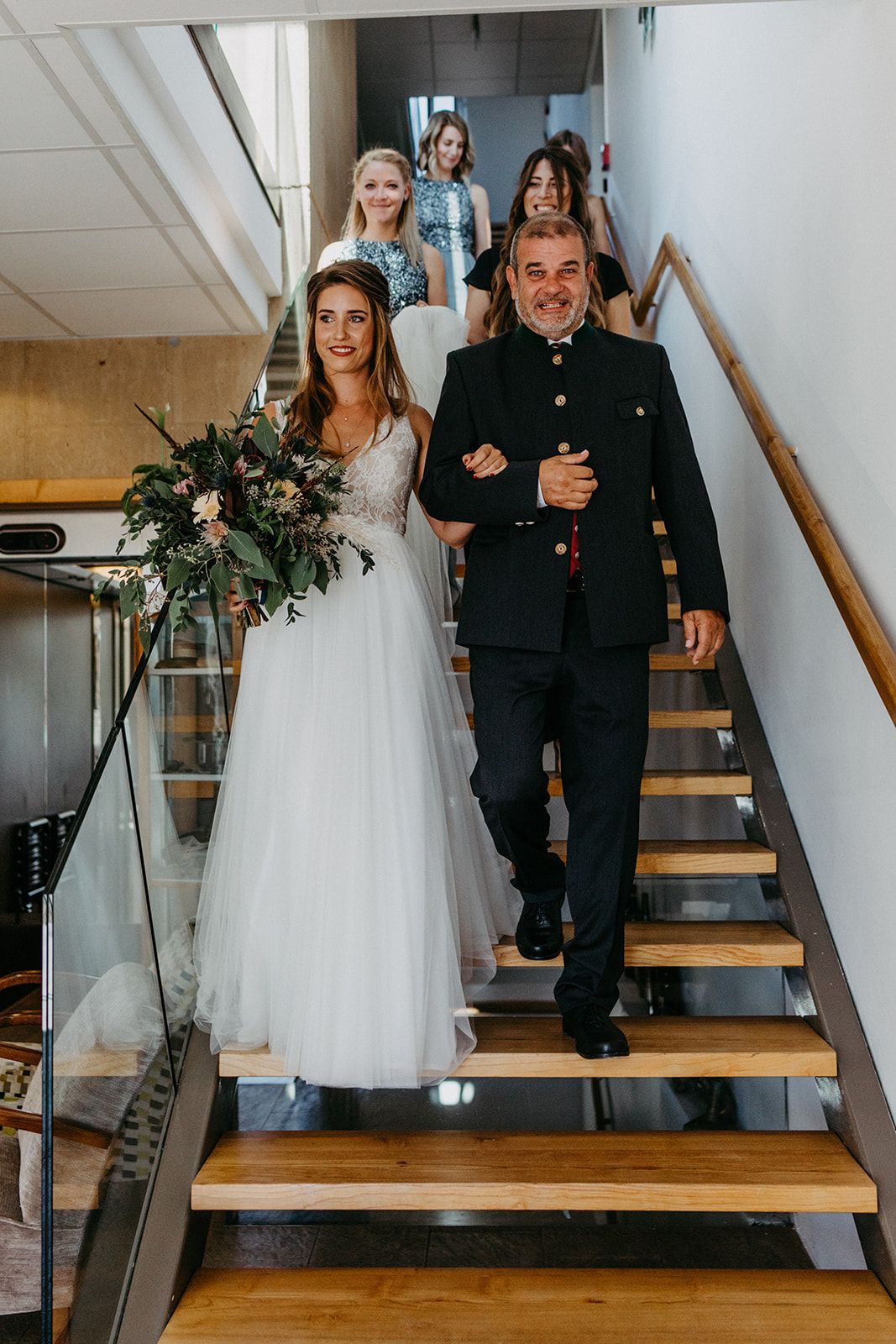 Bride walking down modern staircase with father and bridal party before wedding ceremony.