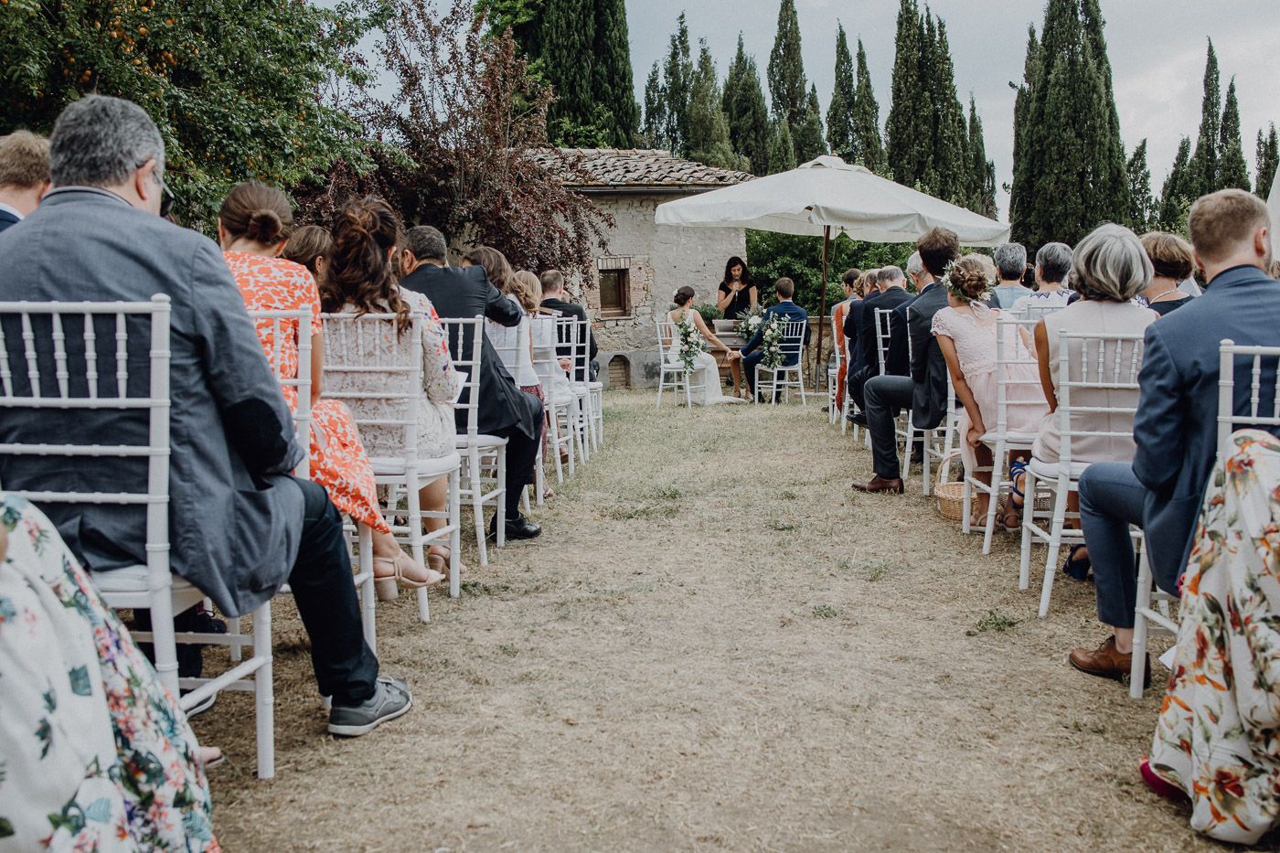 Guests seated in white chairs along garden aisle at elegant outdoor wedding ceremony with cypress trees. Tuscany Villa Catignano
