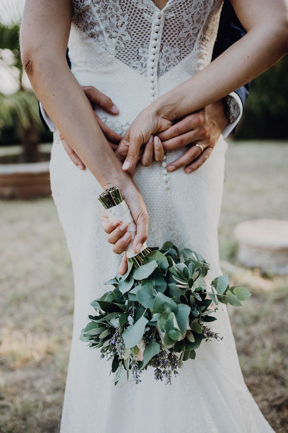 Close-up of bride and groom holding green eucalyptus bouquet with grooms arms wrapped around bride in lace dress. Tuscany Villa Catignano