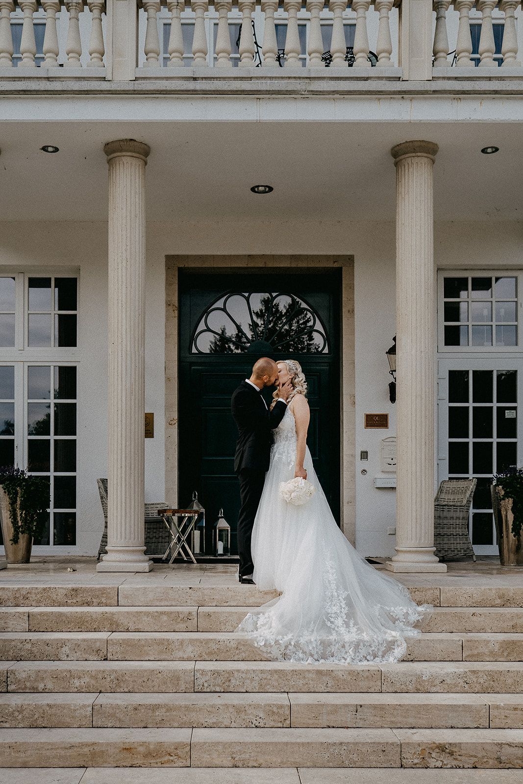 Bride and groom share a kiss on grand steps of an elegant venue with tall white columns.