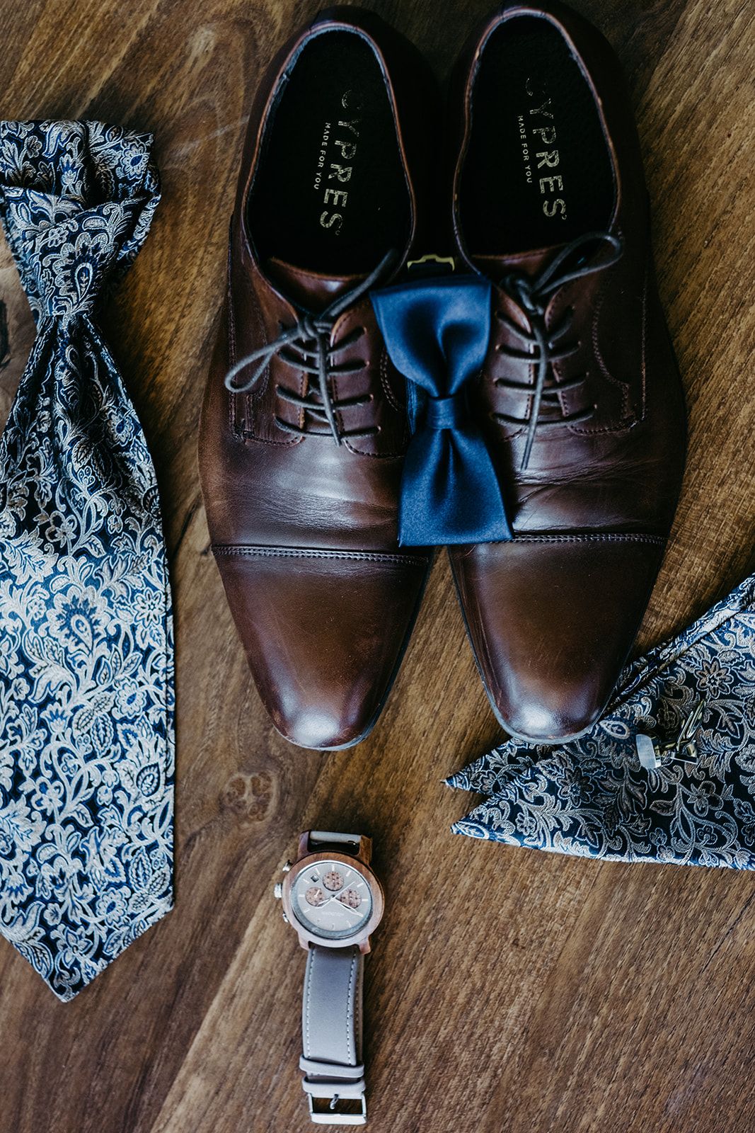Brown leather dress shoes with blue bow tie, paisley tie, watch, and pocket square arranged on wood floor.