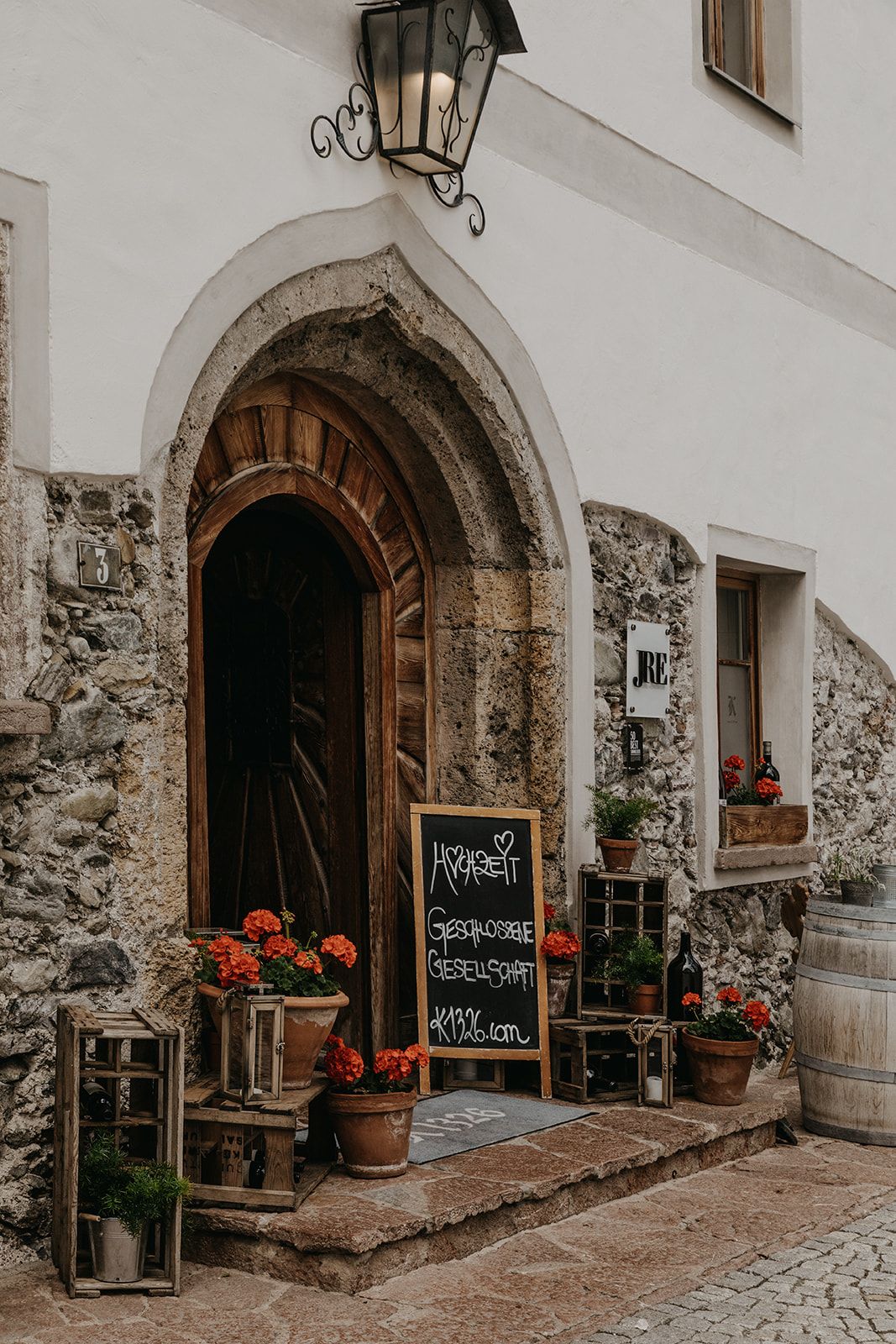 Rustic stone archway entrance of a European restaurant decorated with red geraniums, potted plants, and a chalkboard menu sign. Leogang