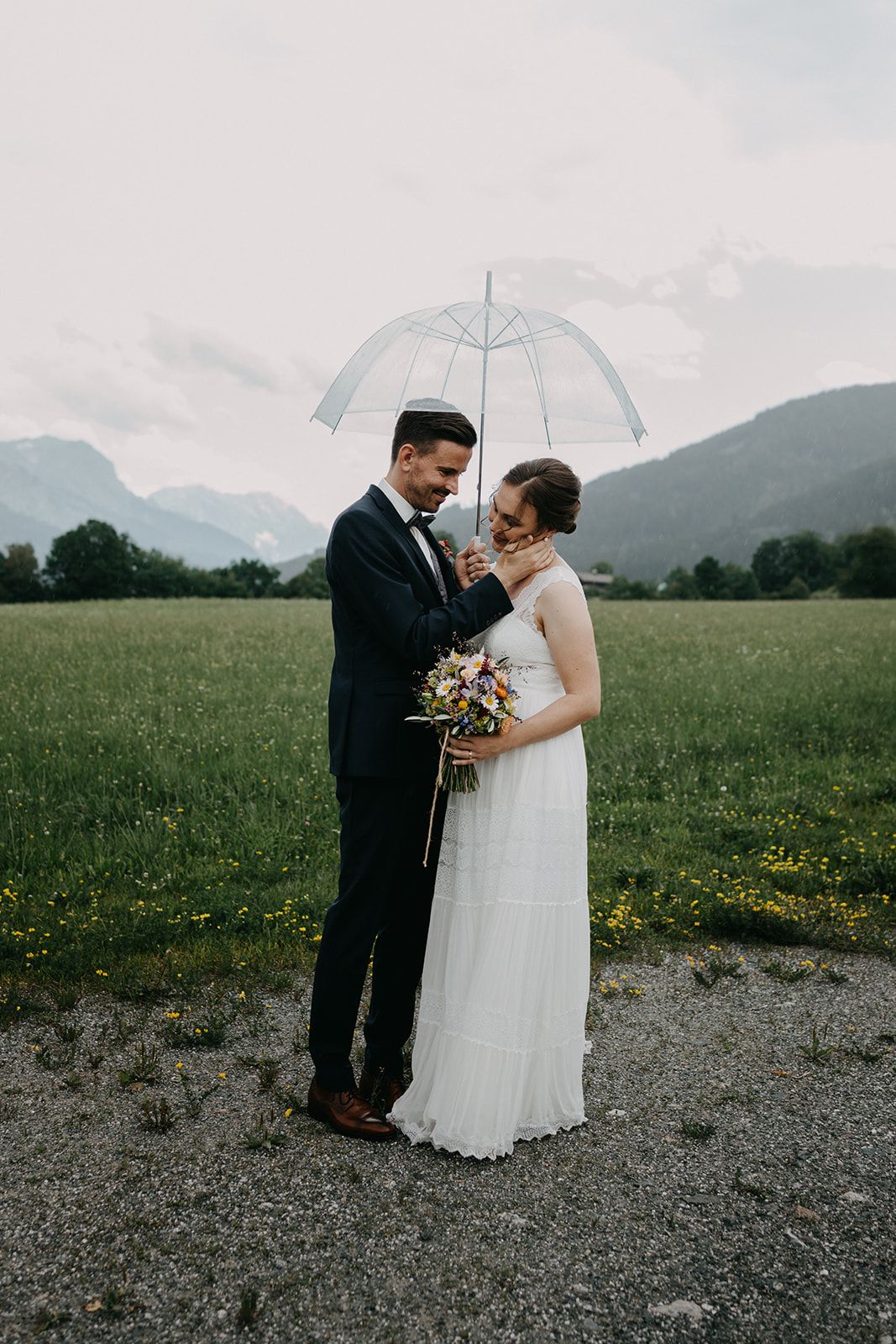 Wedding couple sharing a tender moment under a clear umbrella in a green meadow with mountain views. Leogang Kirchenwirt