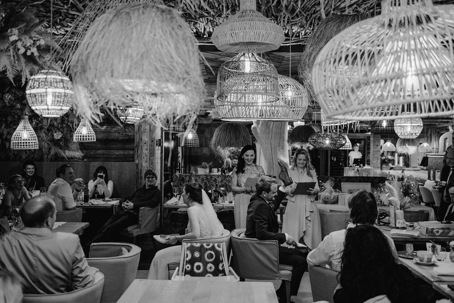 Black and white wedding reception scene with guests dining under decorative wicker pendant lights. Jufenalm