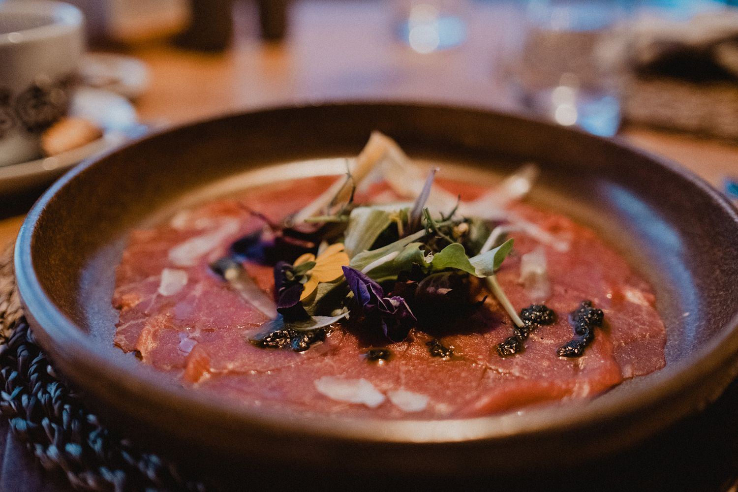 Gourmet carpaccio dish garnished with microgreens and edible flowers served in a rustic ceramic bowl. Jufenalm