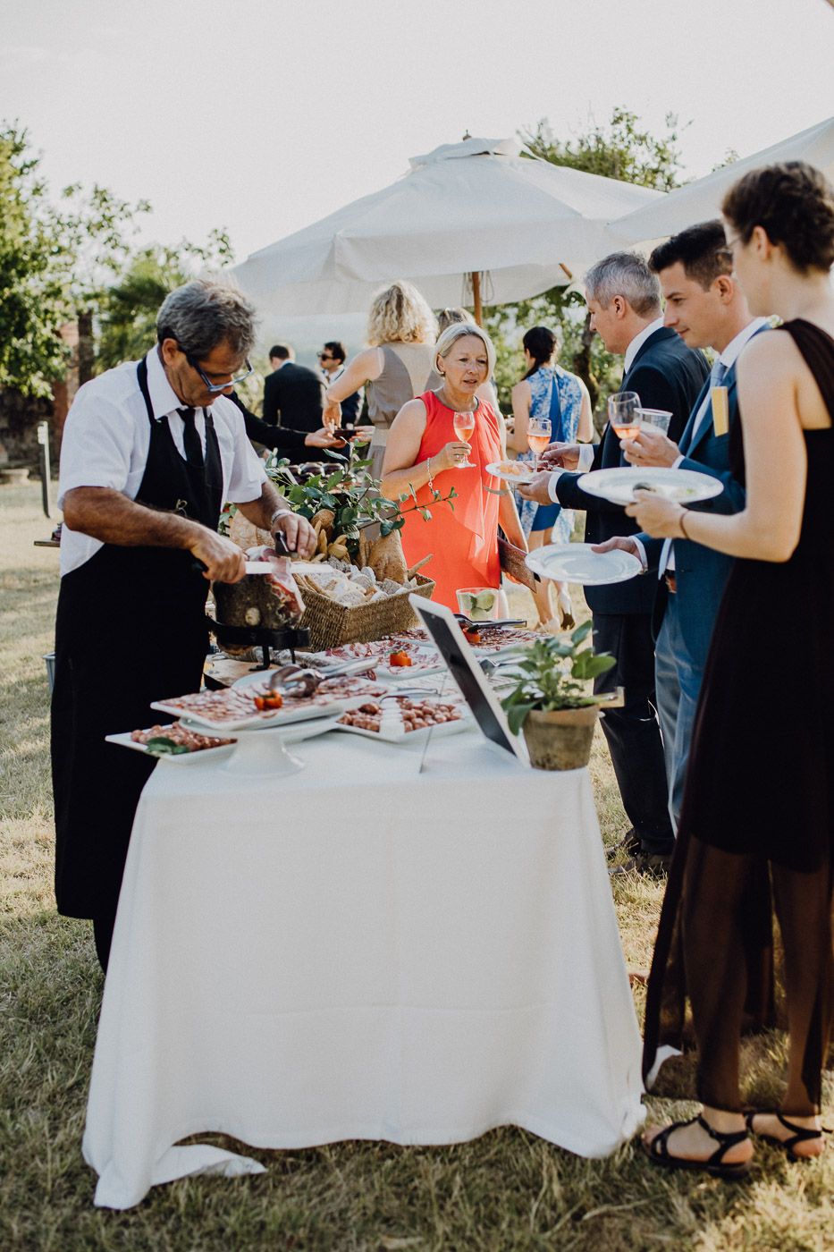 Catered outdoor wedding reception with guests enjoying food stations in a sunny garden setting. Tuscany Villa Catignano