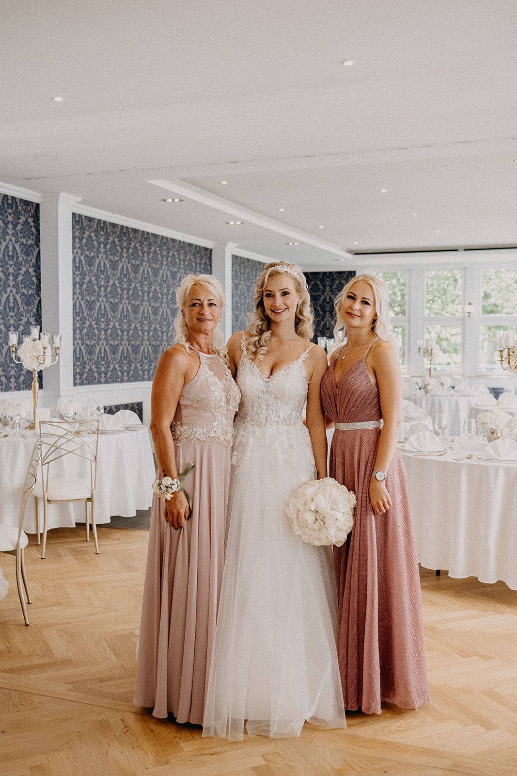 Bride in white lace gown poses with two bridesmaids in dusty rose dresses holding bouquet.