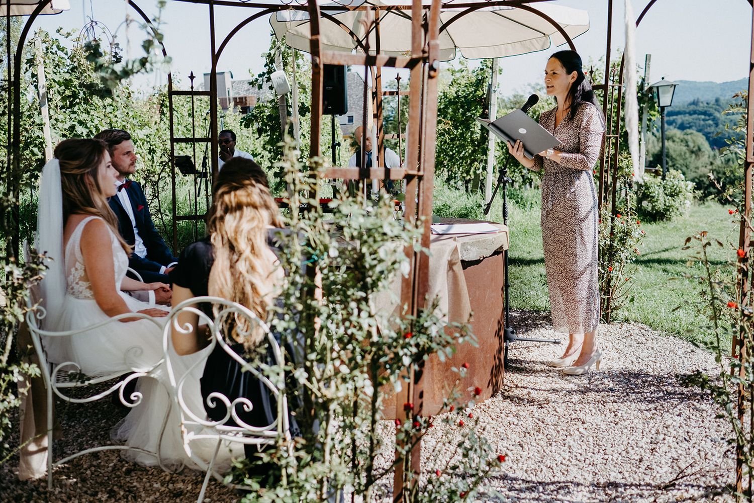 Bride and groom sit during outdoor garden wedding ceremony as officiant reads from a book.