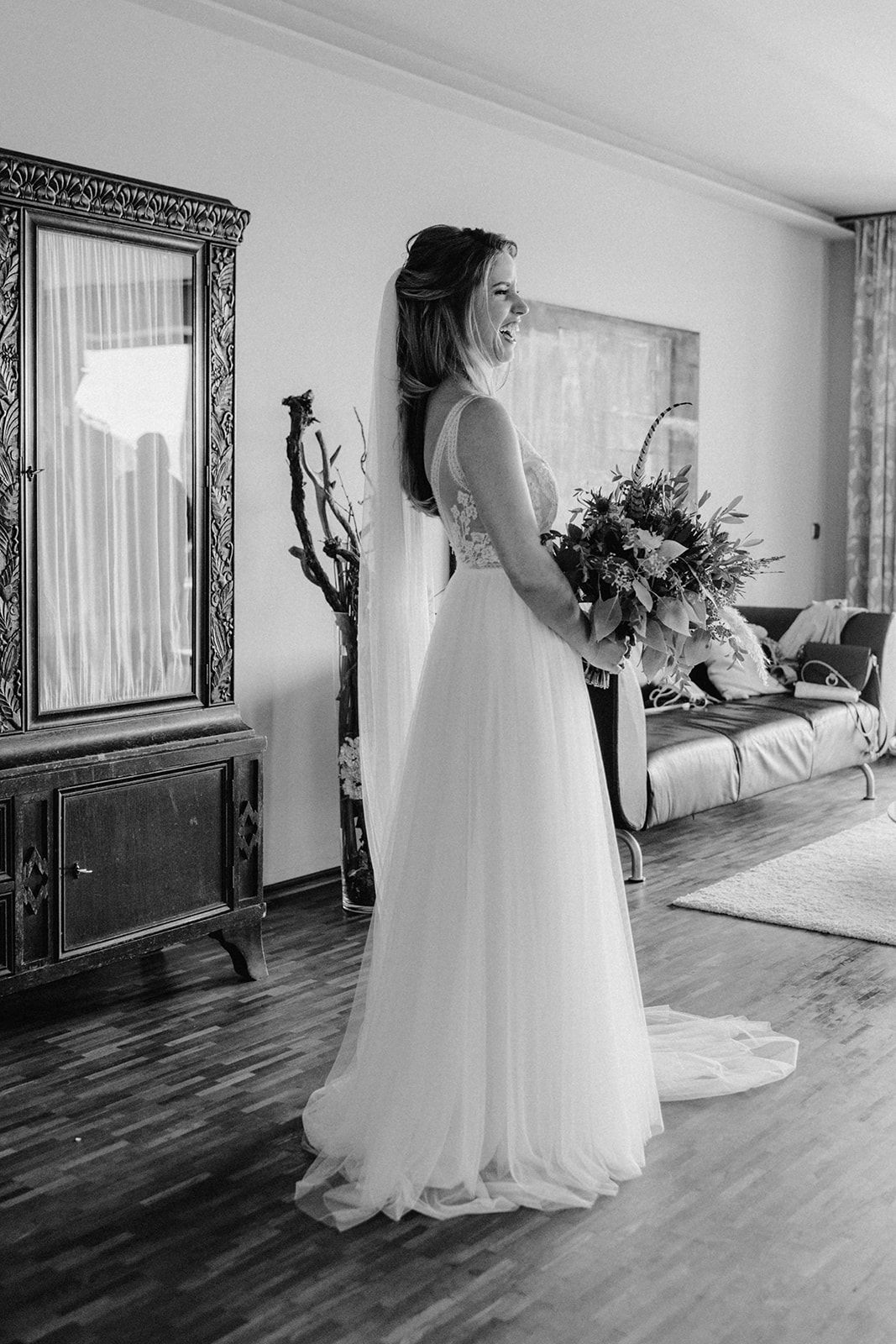 Full length black and white portrait of bride in flowing wedding gown holding bouquet indoors.