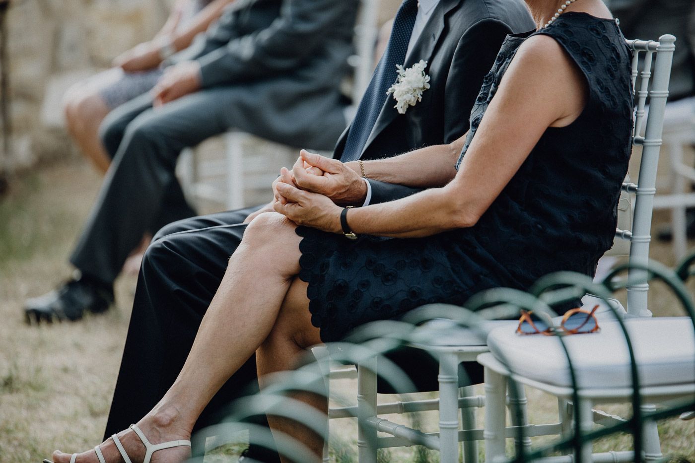 Wedding guest in navy dress holds hands with companion while seated at outdoor ceremony. Tuscany Villa Catignano