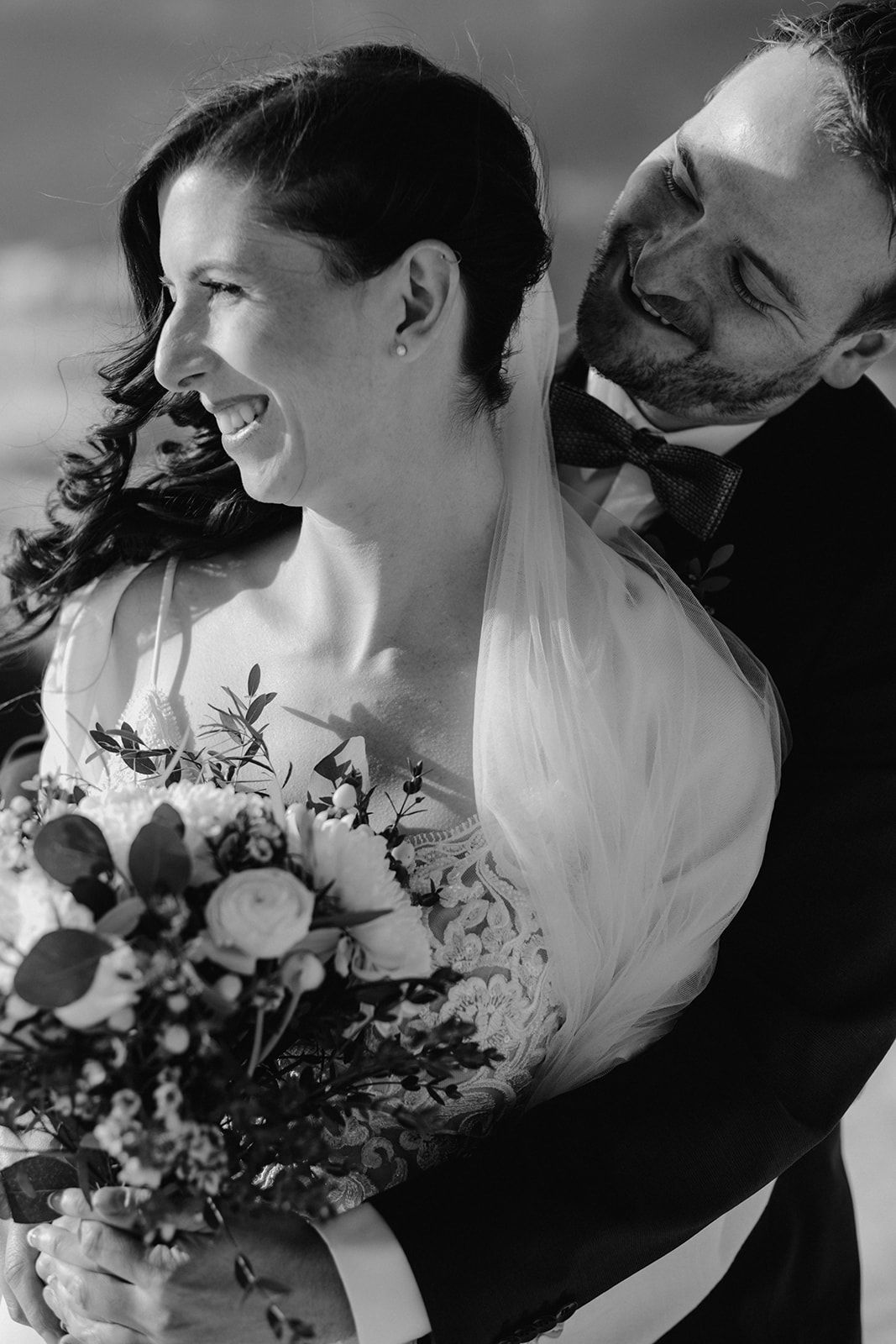 Black and white portrait of laughing bride with bouquet embraced by smiling groom in bow tie at winter wedding. Jufenalm