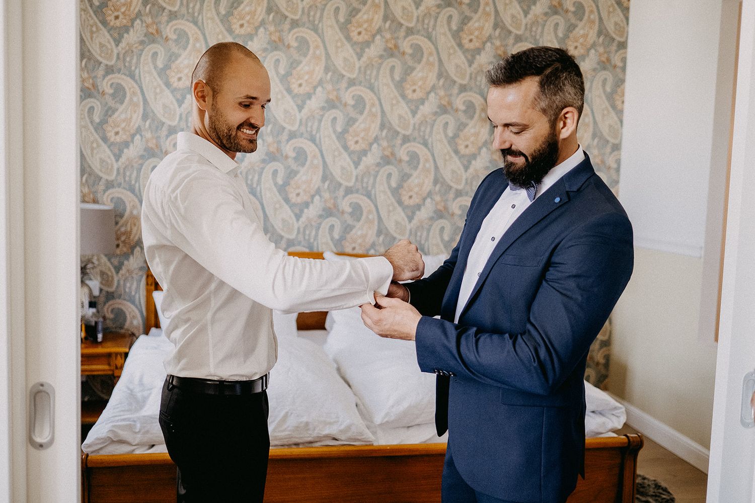Two men in formal wedding attire helping each other with cufflinks in a decorated hotel room.