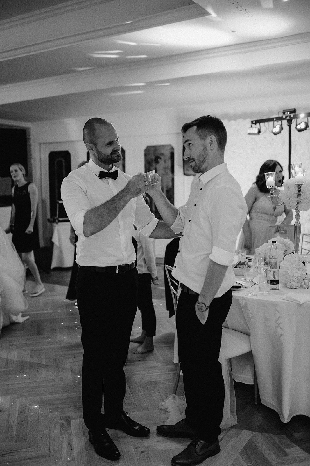 Two men in formal attire clinking glasses at a wedding reception in black and white photo.