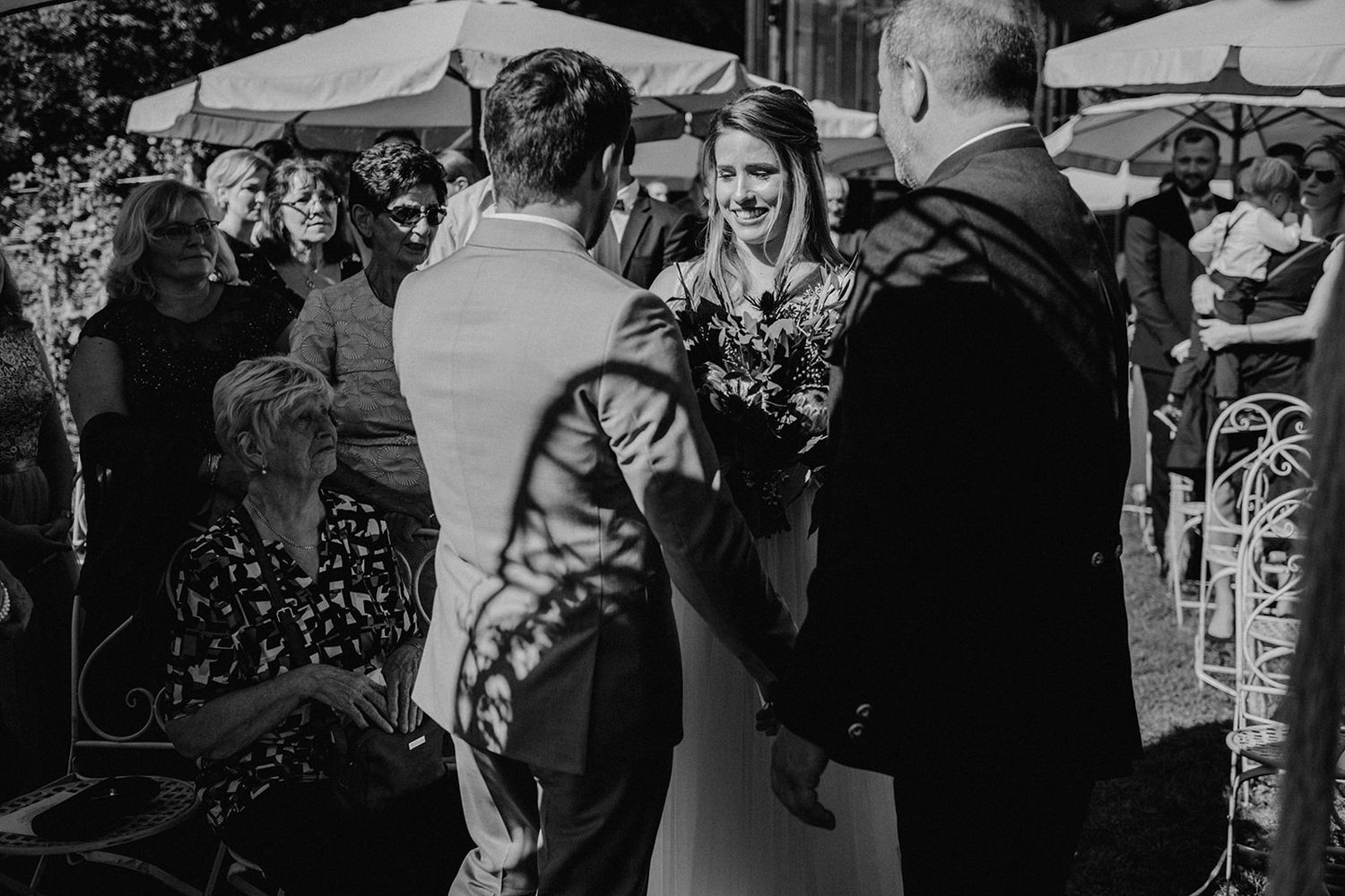 Black and white photo of bride smiling as groom takes her hand at outdoor wedding ceremony.