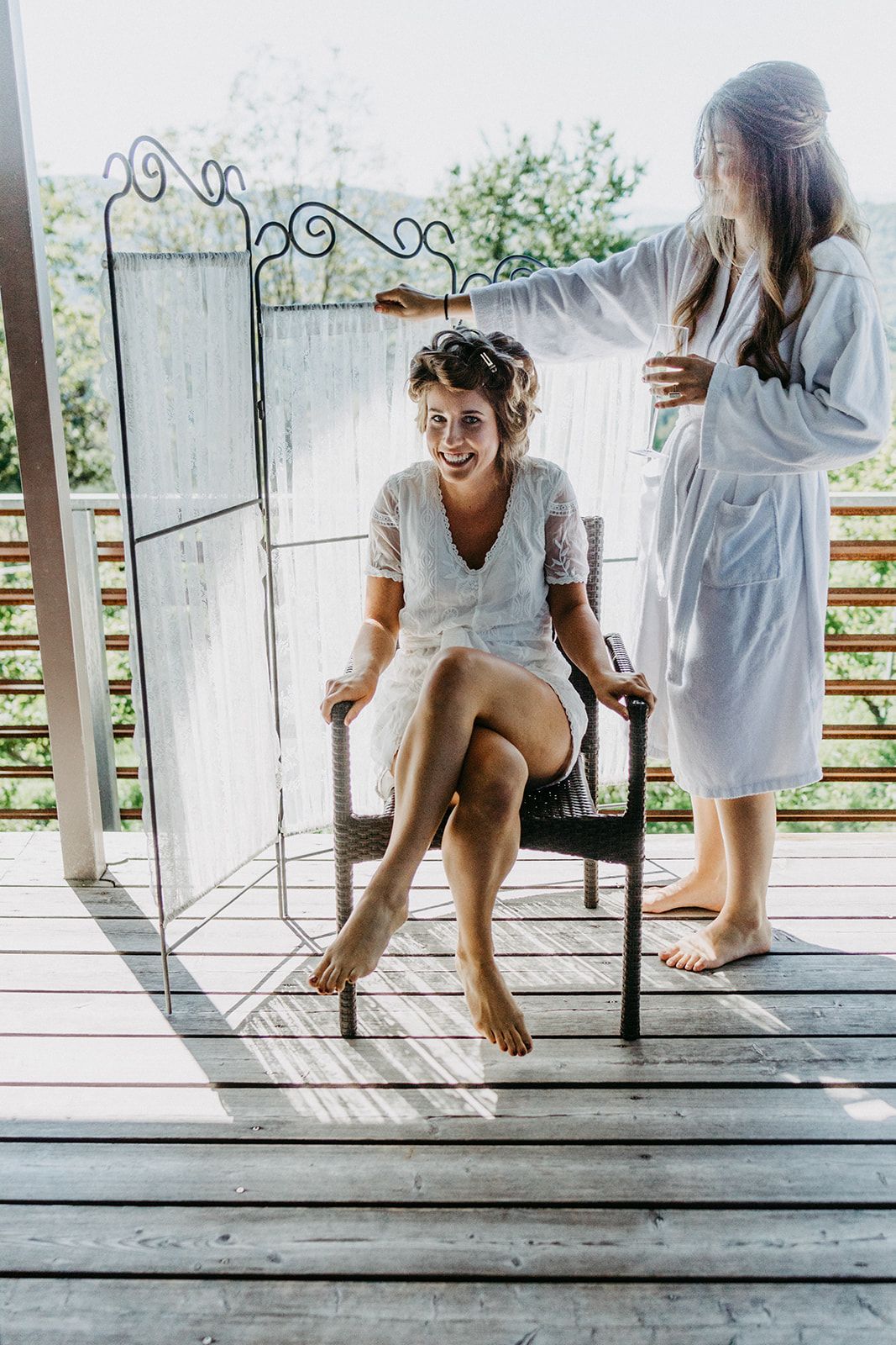Bride sitting in chair on wooden deck while bridesmaid styles her hair outdoors.
