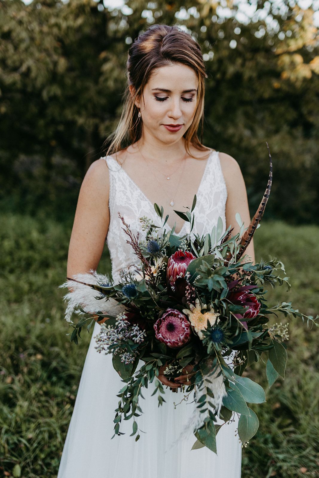 Bride in lace wedding dress holding a lush boho bouquet with protea, eucalyptus, and feathers outdoors.