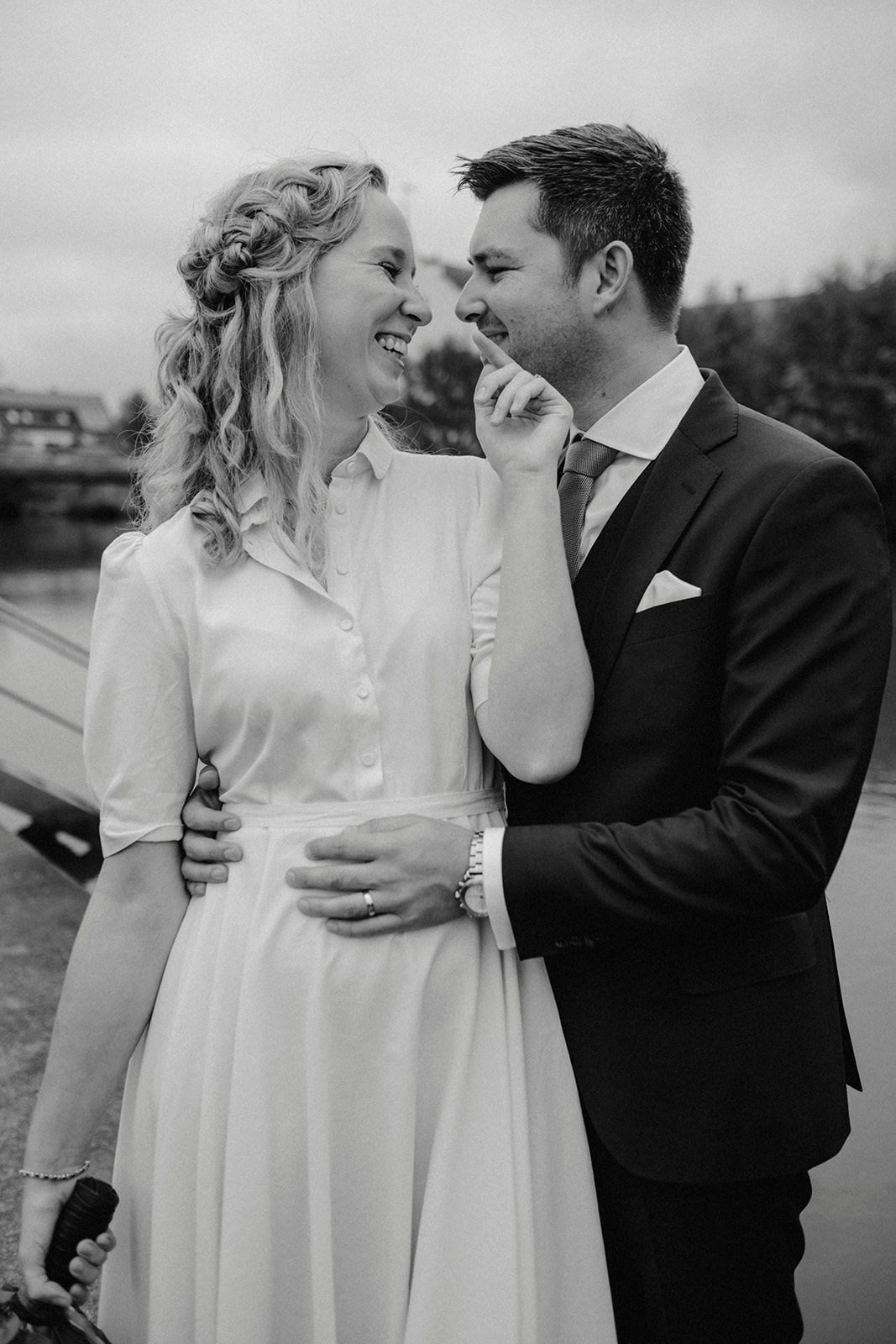 Black and white portrait of smiling bride and groom sharing intimate moment outdoors near waterway.