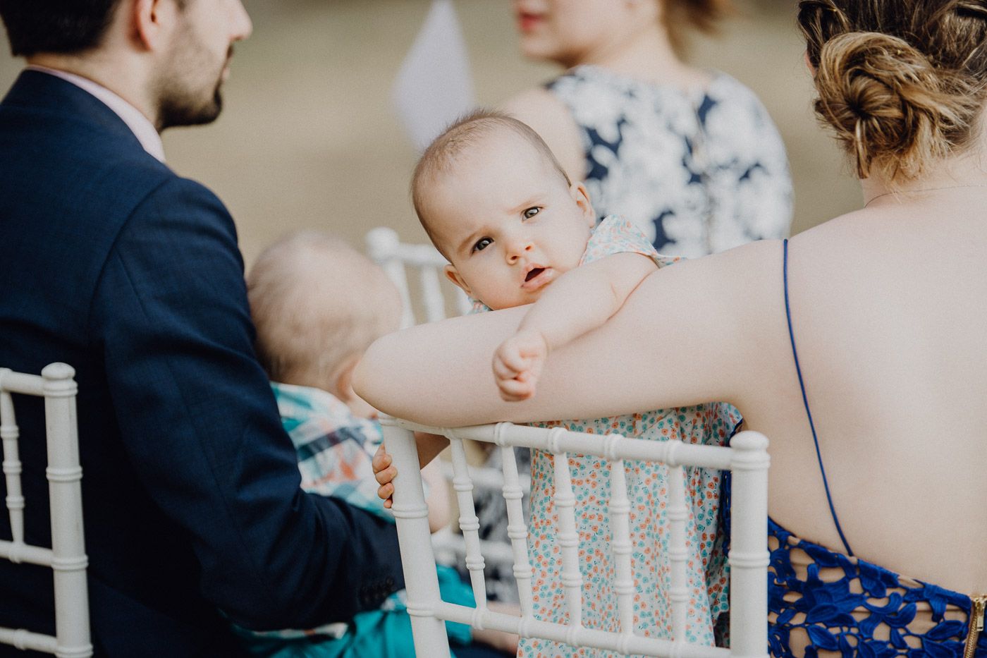 Adorable baby in blue outfit looks around curiously while being held at outdoor wedding ceremony. Tuscany Villa Catignano
