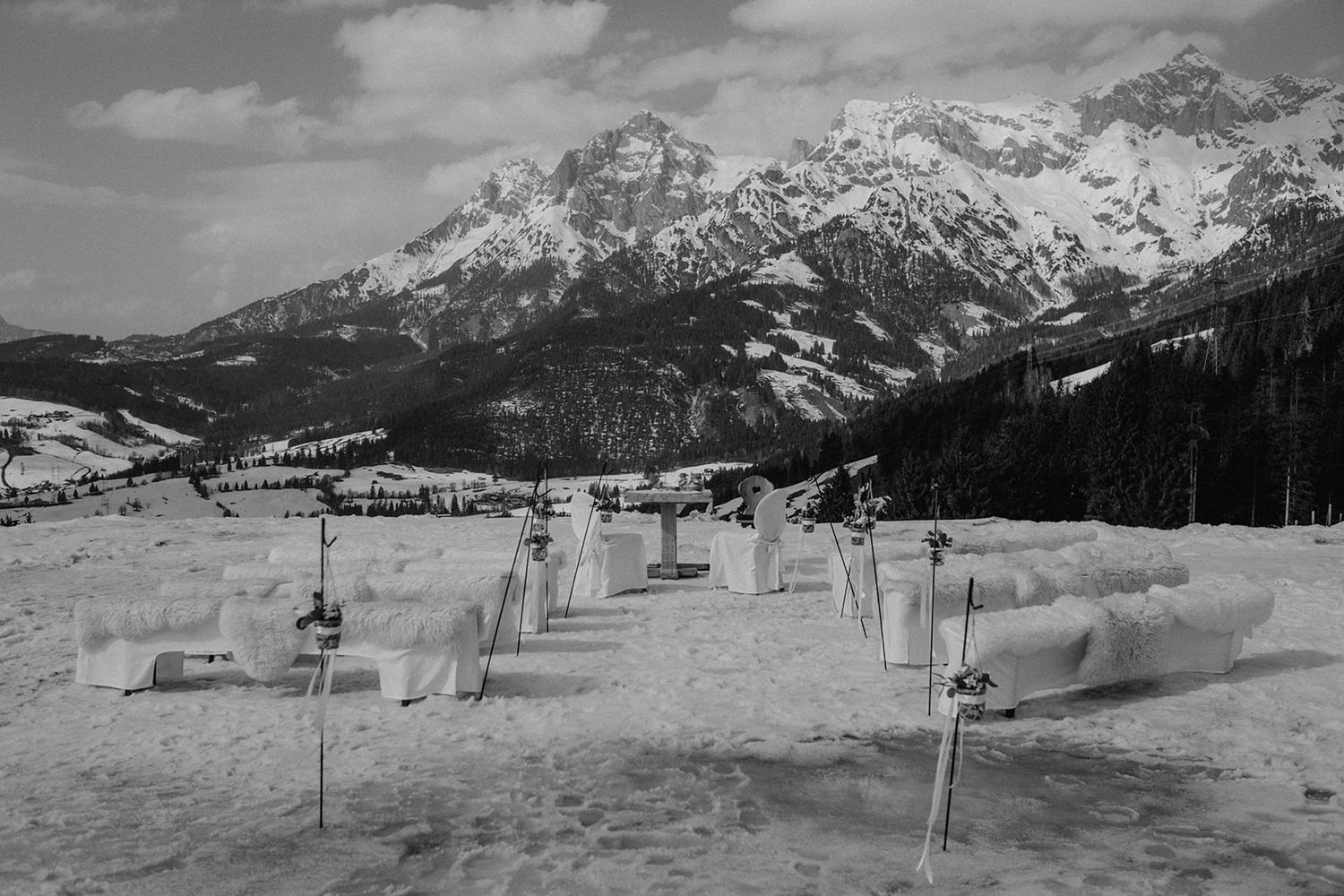 Black and white landscape of snow-covered Alpine mountains with ice sculptures arranged in a snowy field below. Jufenalm