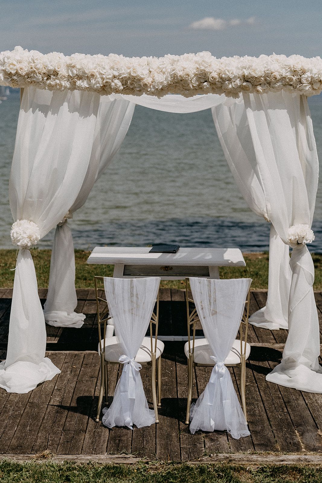 Elegant waterfront wedding ceremony arch draped in white fabric with floral accents and two decorated chairs.