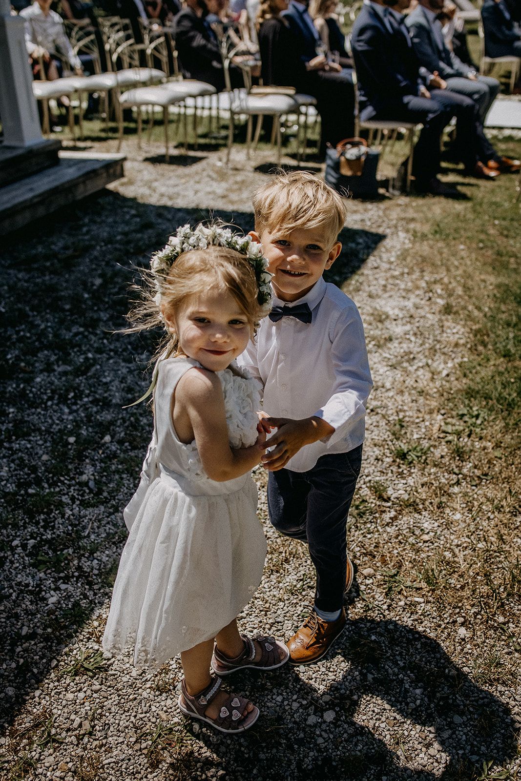 Two adorable flower girl and ring bearer holding hands at an outdoor wedding ceremony.