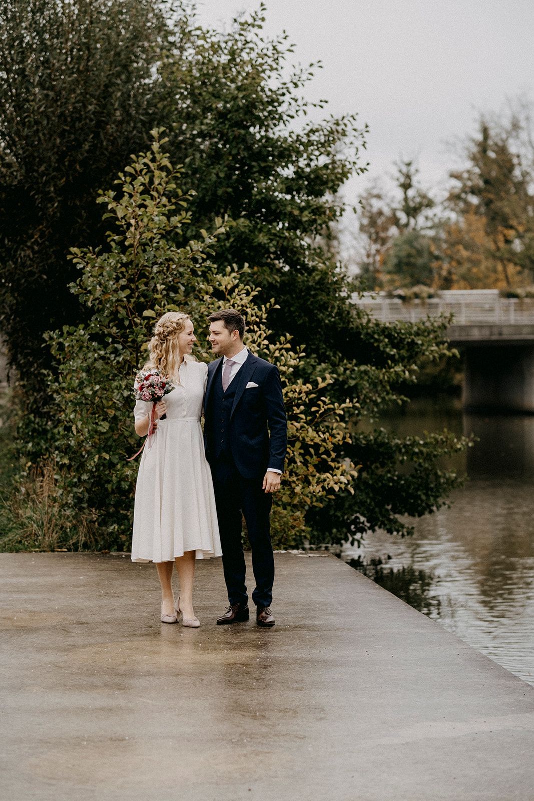 Bride and groom in wedding attire walking together on a wet pathway beside a river surrounded by lush greenery.