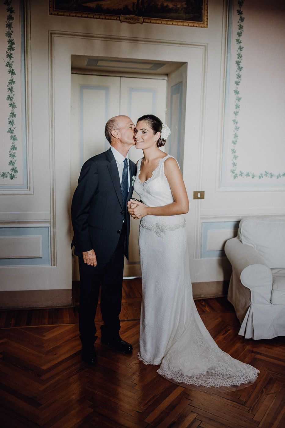 Bride in white gown receives kiss on cheek from older man in dark suit at elegant wedding venue. Tuscany Villa Catignano
