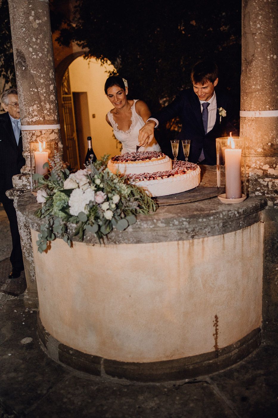 Bride and groom cutting wedding cake on stone table with candles and floral arrangements at elegant outdoor venue. Tuscany Villa Catignano