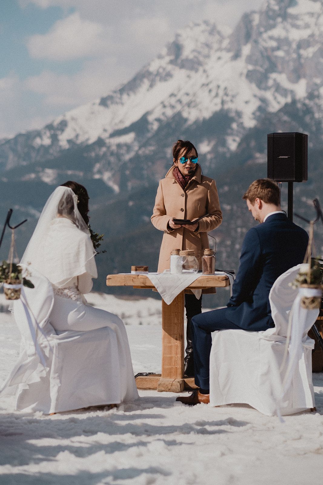 Outdoor winter wedding ceremony with officiant reading vows before snow-capped mountain peaks.