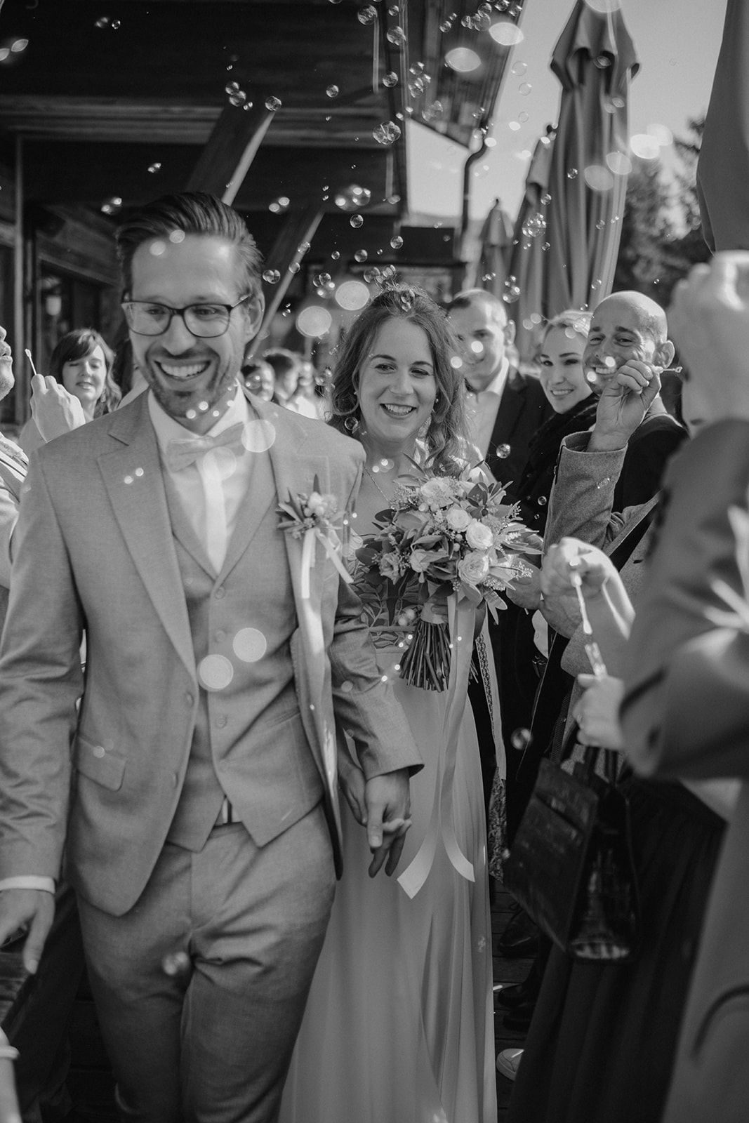 Smiling newlyweds walking through bubbles with wedding guests, black and white.