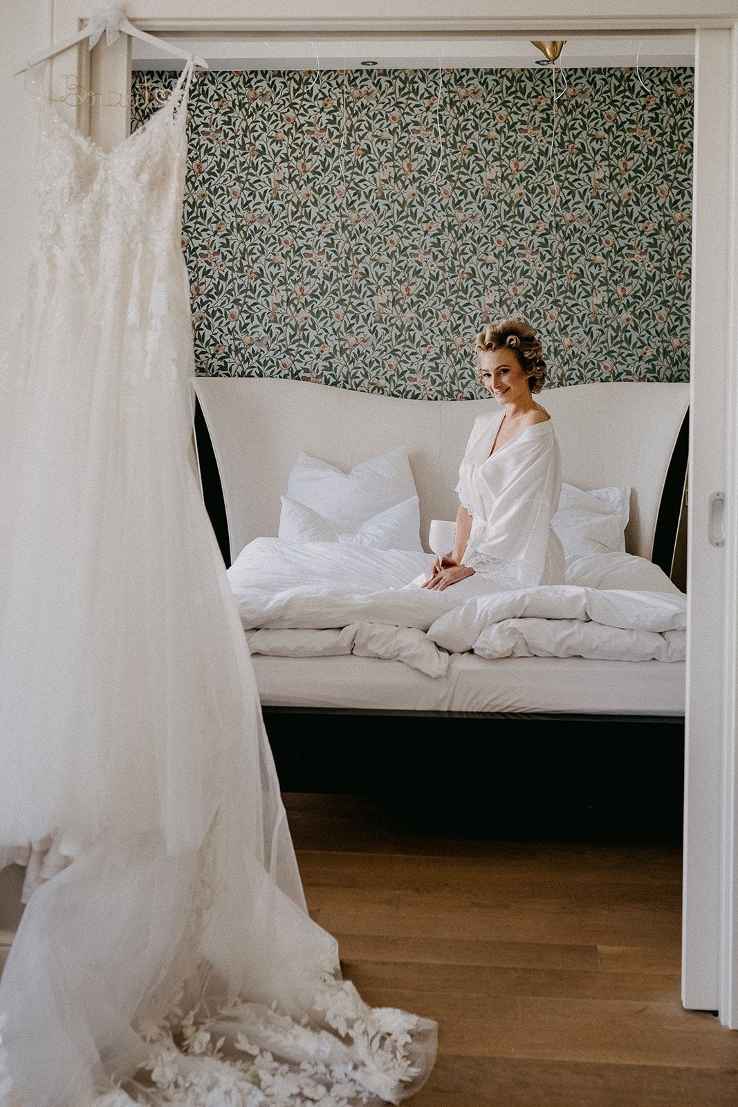Smiling bride in white robe sitting on bed with wedding dress hanging in foreground of elegant room.