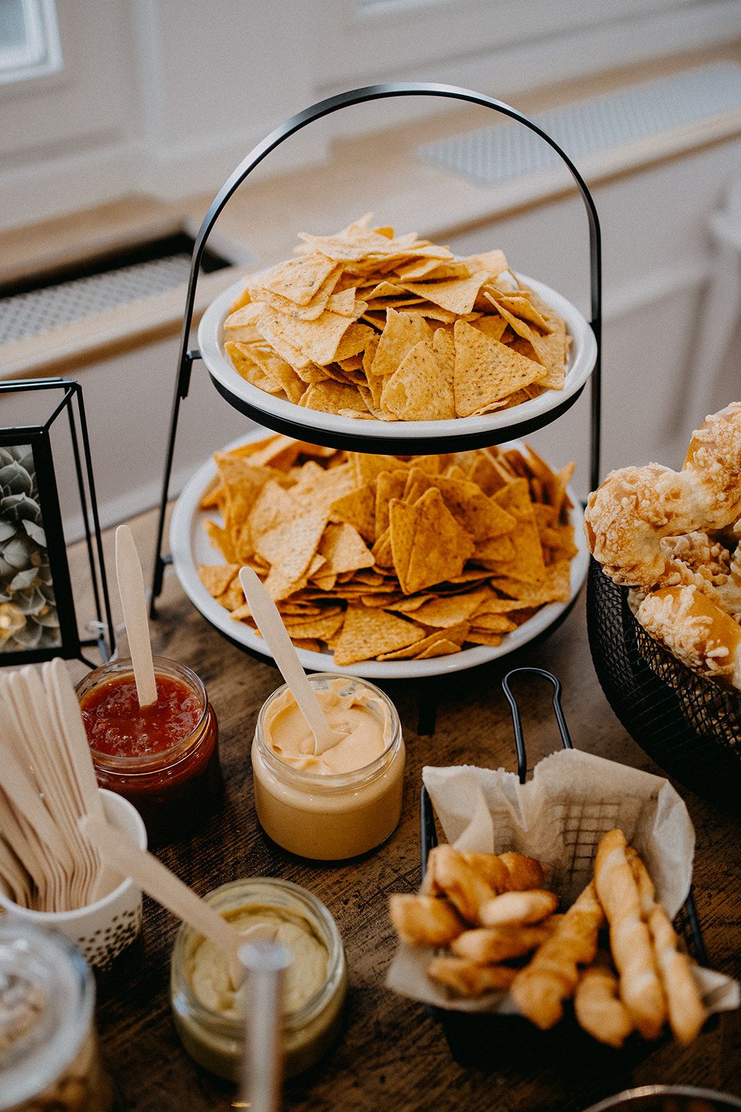 Two-tiered serving stand with tortilla chips and dipping sauces at wedding snack station.