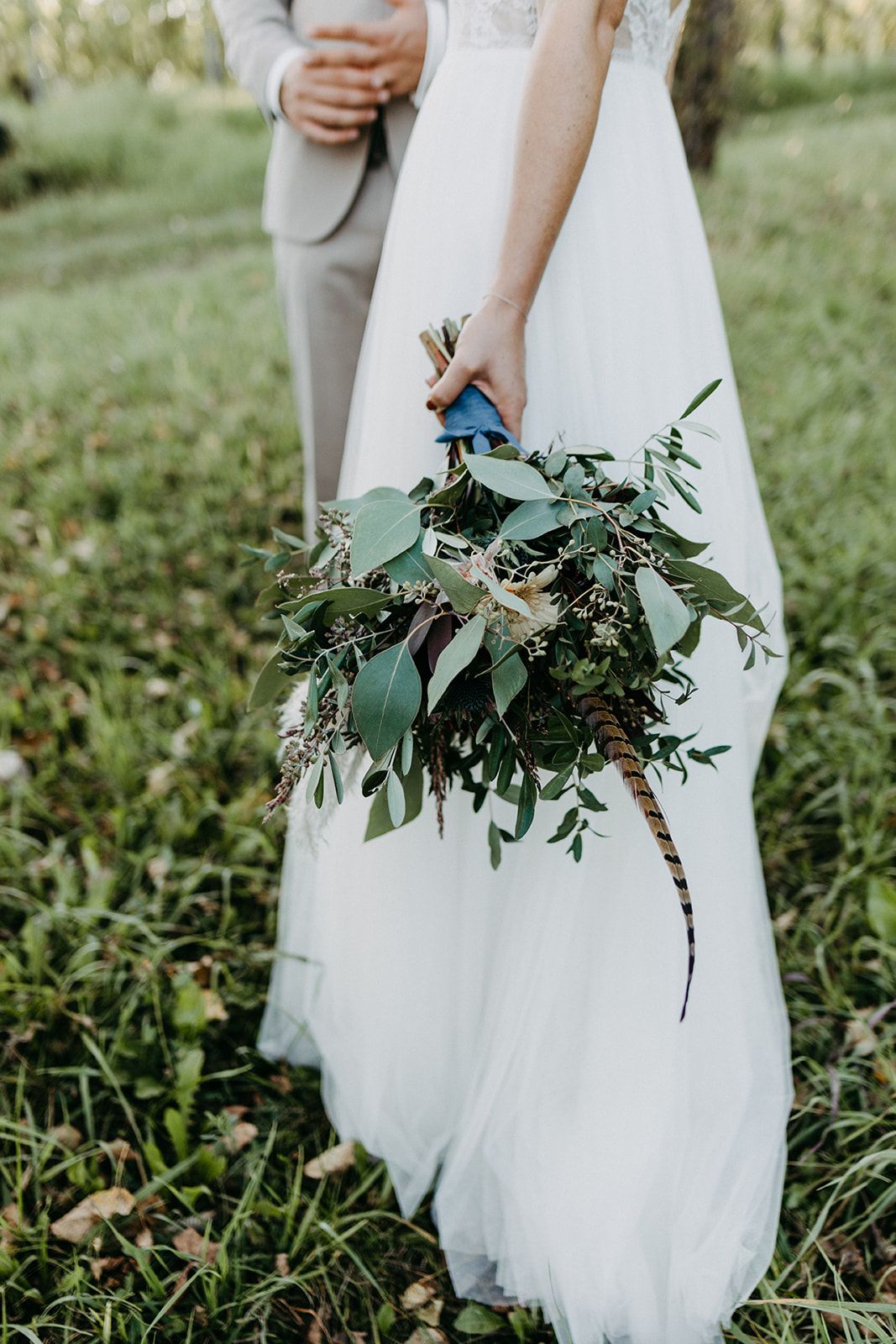 Bride in white gown holding a greenery-filled bouquet with feathers while standing with groom in a lush garden.
