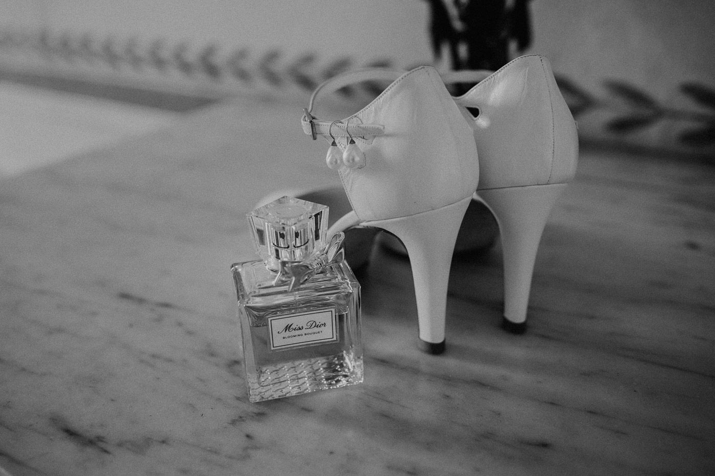 Black and white photo of Miss Dior perfume bottle beside white bridal heels on marble. Tuscany Villa Catignano