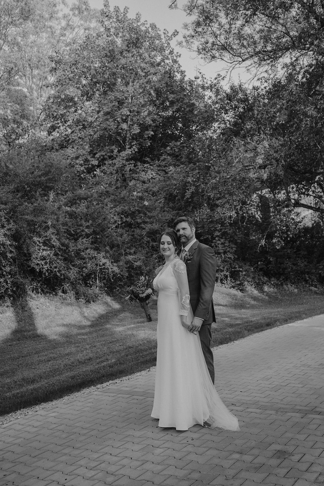 Black and white portrait of a bride and groom holding hands on a tree-lined pathway.