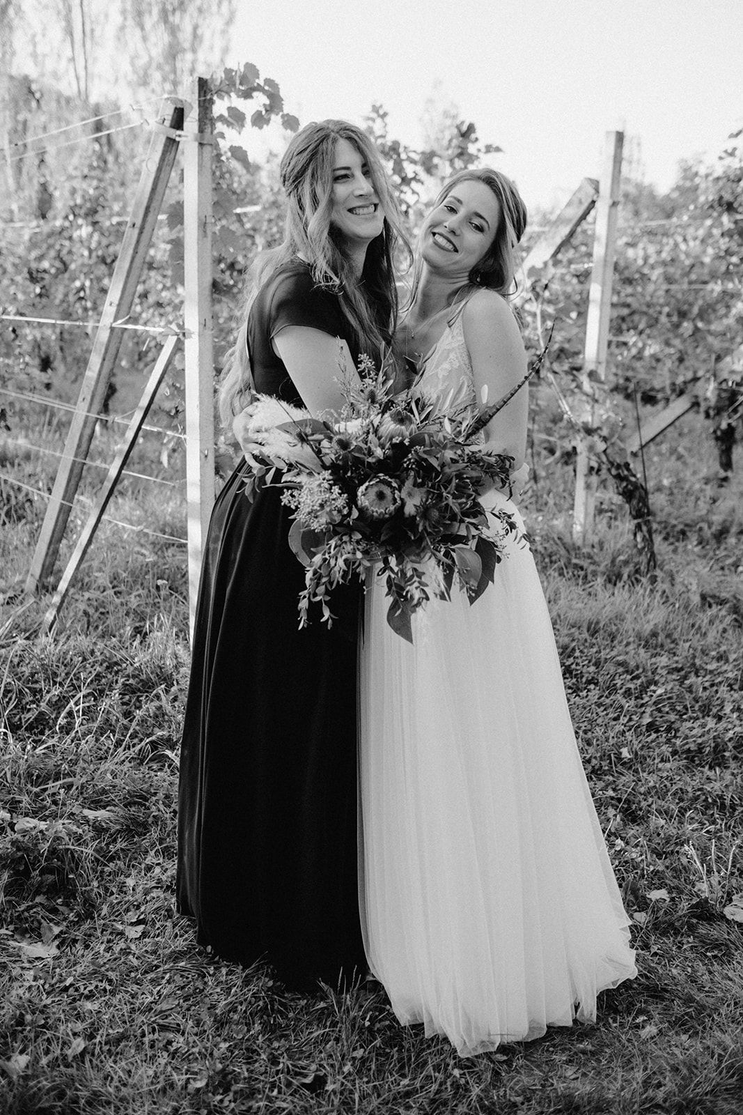 Black and white photo of bride in white gown and bridesmaid embracing while holding a floral bouquet in a vineyard.
