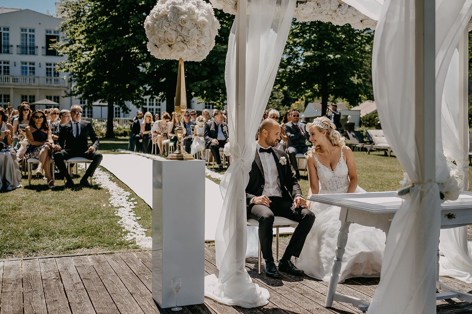 Bride and groom sit together under white draped floral wedding arch during outdoor ceremony. Hideaway Starnberger See