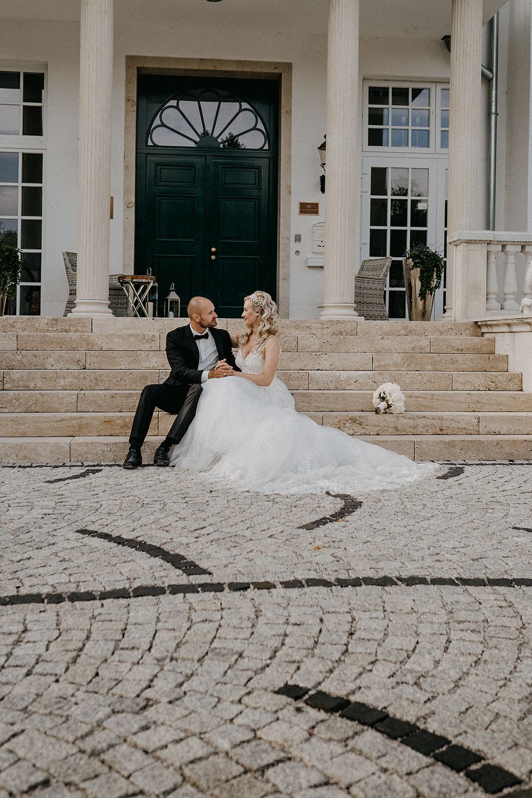 Bride and groom sit together on cobblestone steps of an elegant venue, laughing joyfully.
