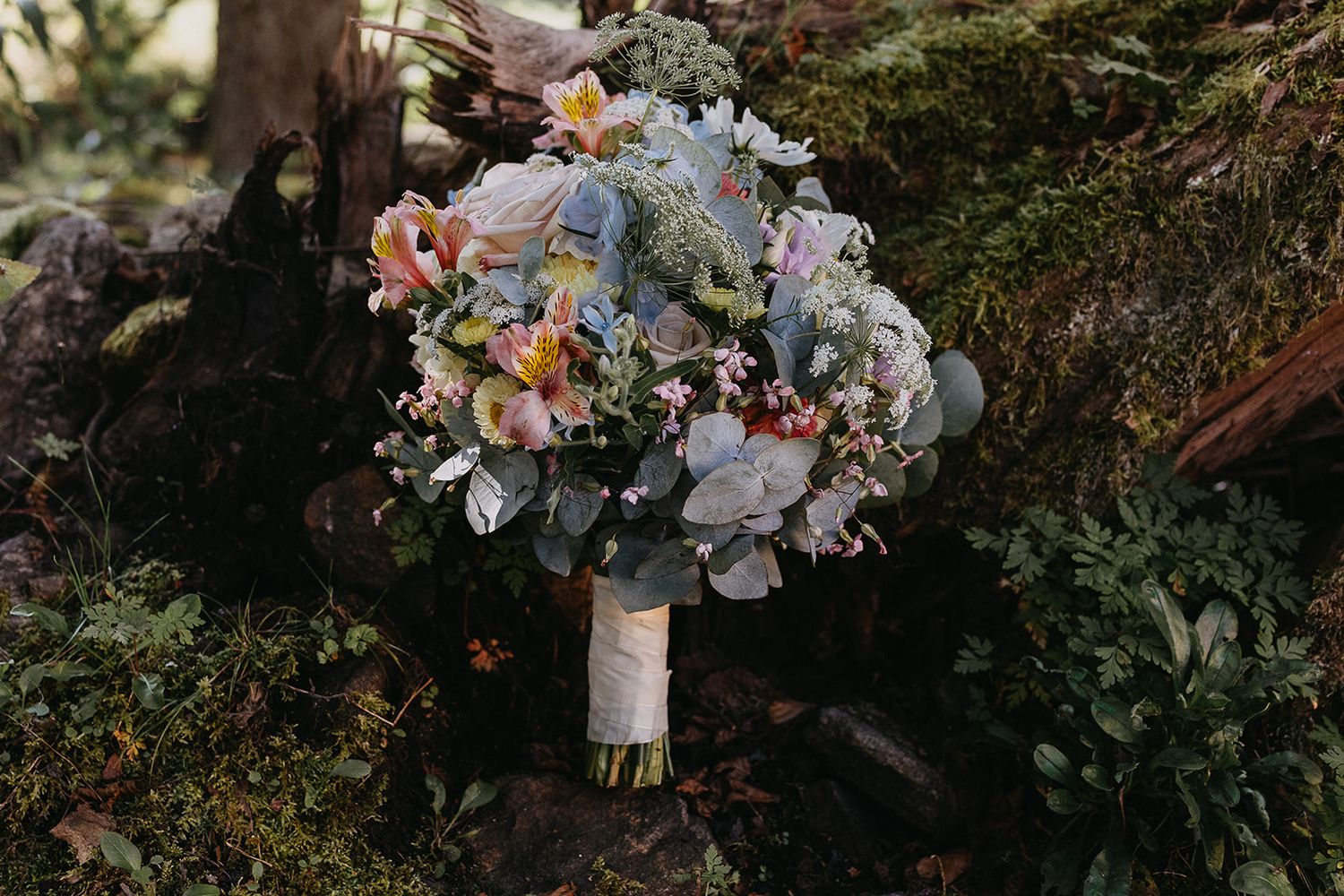 Romantic bridal bouquet with pink, blue, and white flowers resting on a mossy log.