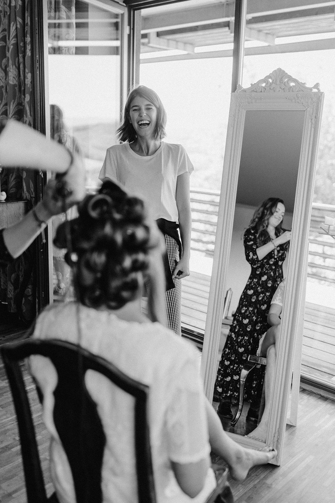 Black and white photo of bride getting hair done while bridesmaid laughs near ornate mirror.