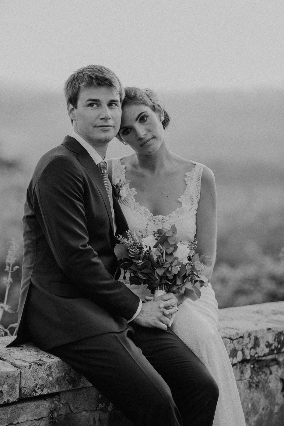 Black and white portrait of bride and groom sitting on stone wall, bride holding bouquet. Tuscany Villa Catignano