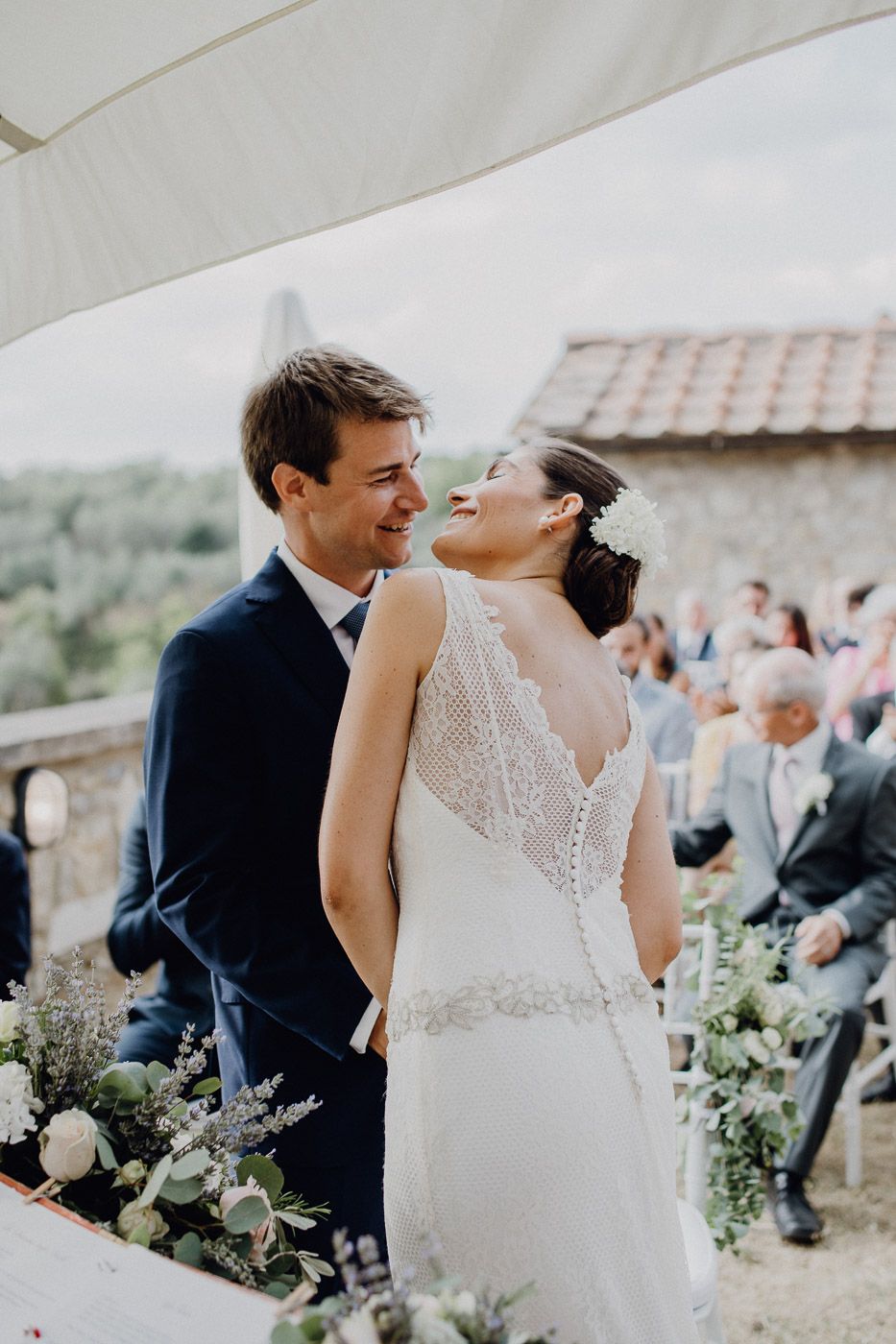 Joyful bride and groom share a tender moment together at outdoor wedding ceremony with guests seated behind them. Tuscany Villa Catignano