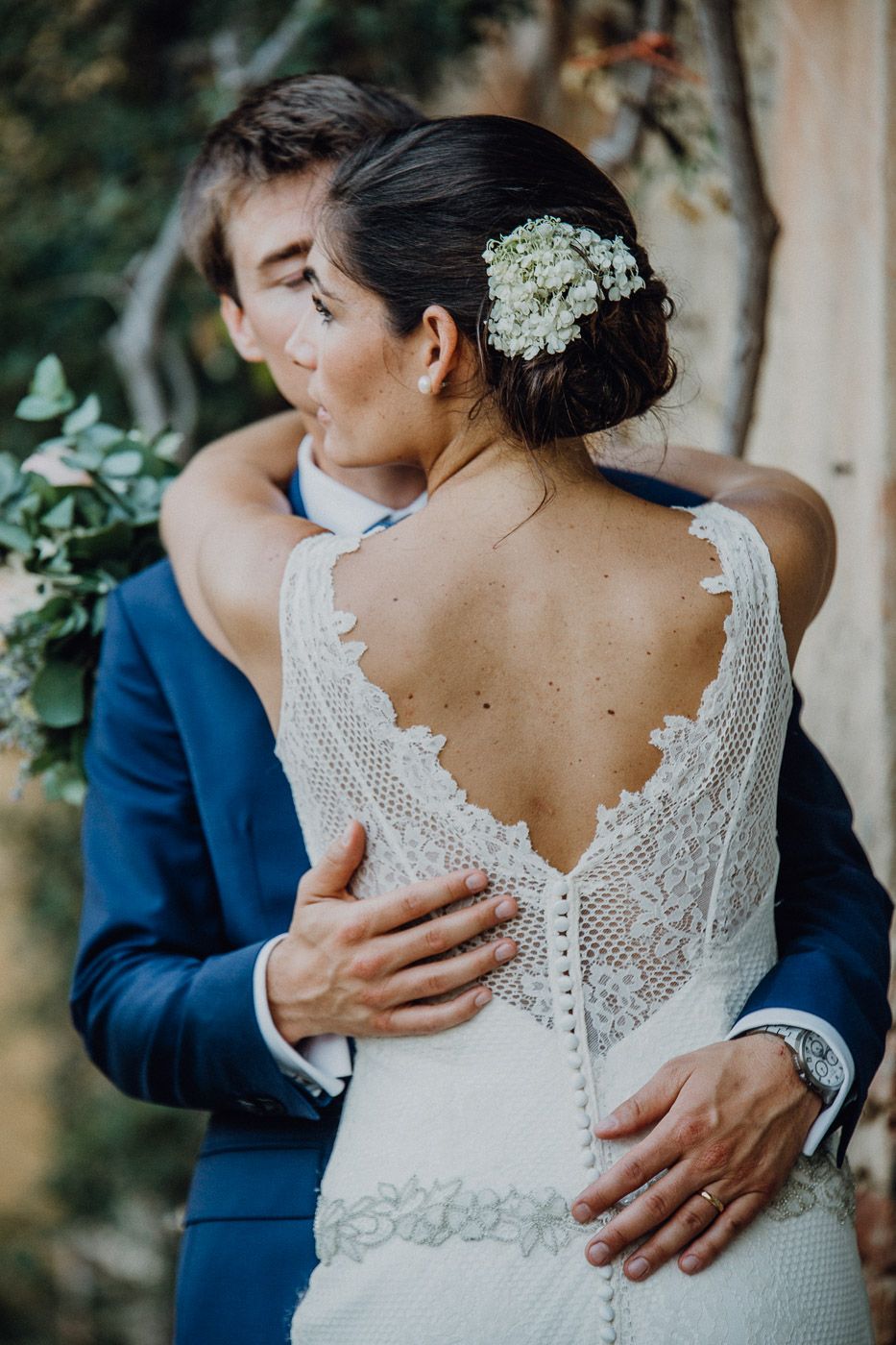 Bride in lace backless wedding gown with floral hair accessory embraced by groom outdoors.