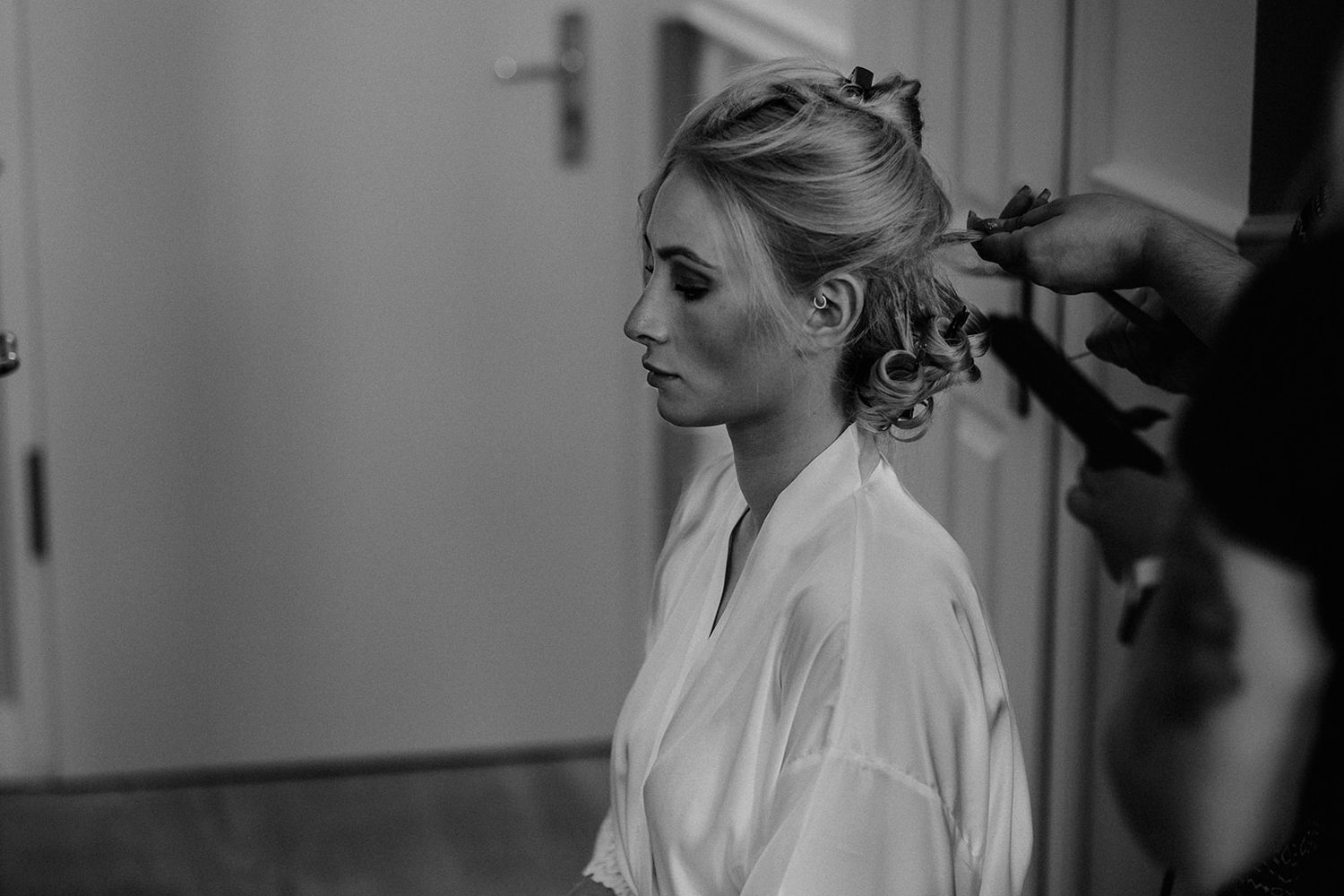 Bride in white silk robe getting hair styled in elegant updo during wedding preparations, black and white photo.