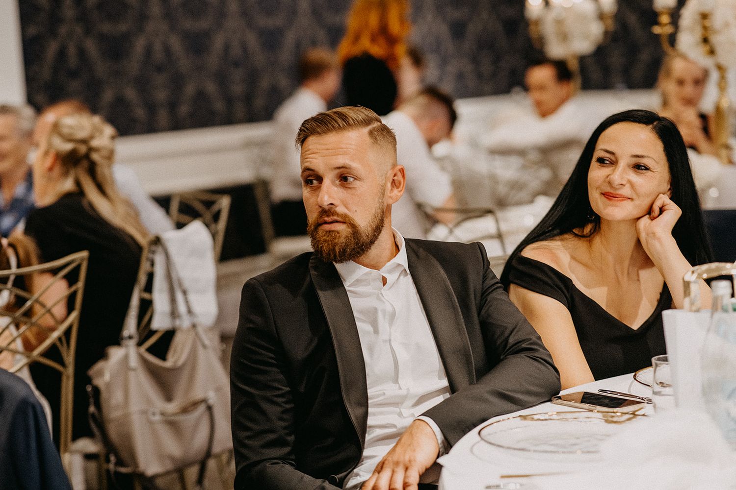 Stylish wedding guests, a bearded man in a suit and woman in black dress, seated at a reception dinner table.