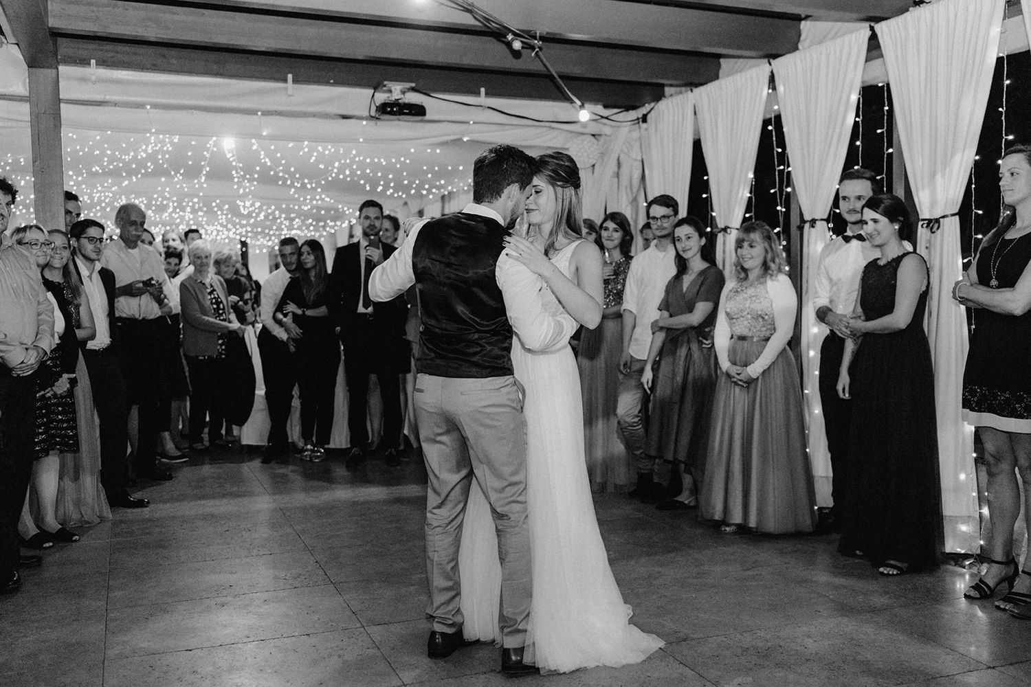 Newlyweds share an intimate slow first dance while wedding guests watch and celebrate around them.