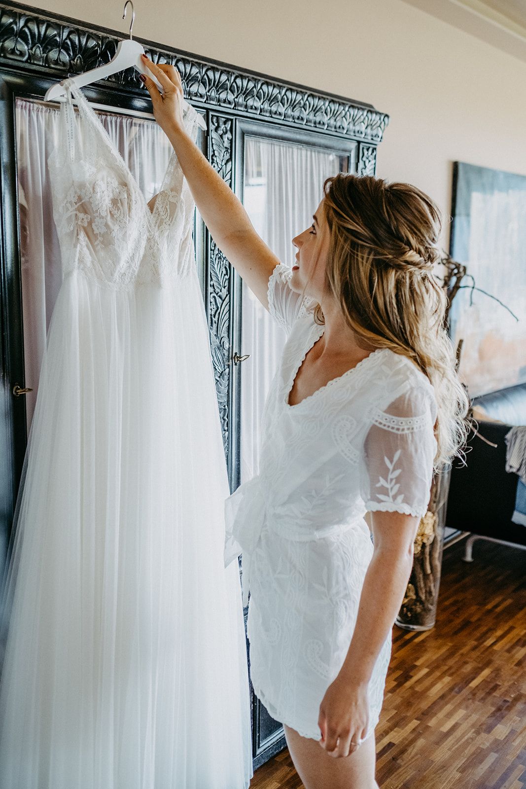 Bride in white robe reaching up to touch her wedding dress on hanger.